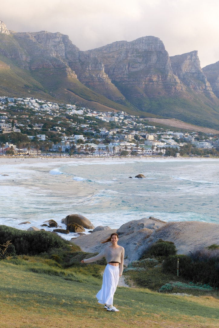 A woman in a white skirt on a grassy headland above the sea, with mountains and a town behind
