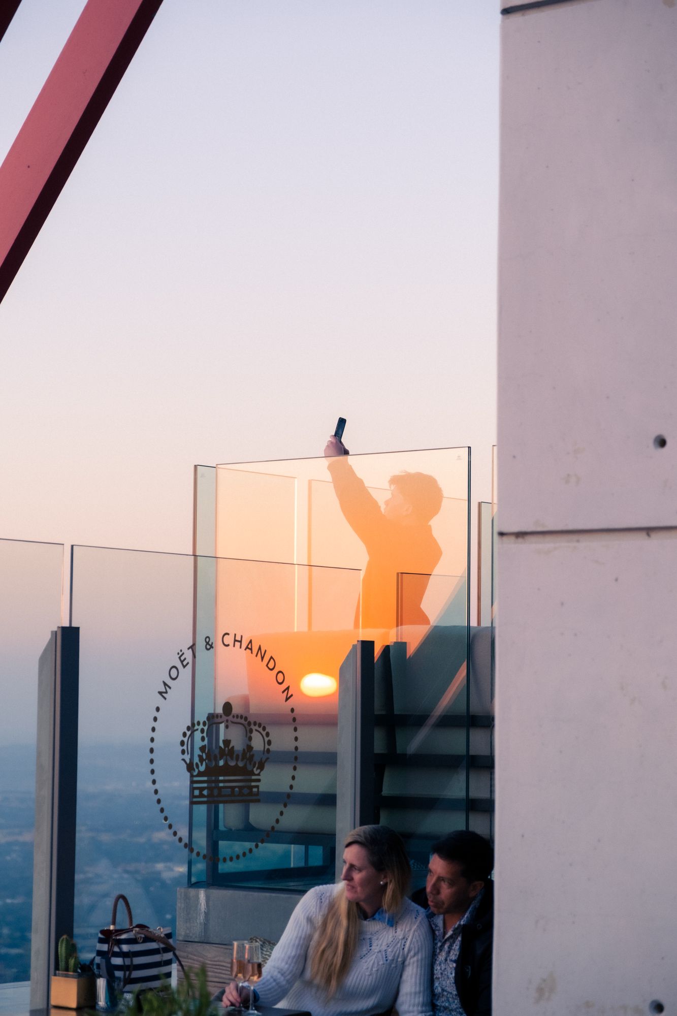 A silhouette taking a photo behind glass at a rooftop bar at sunset